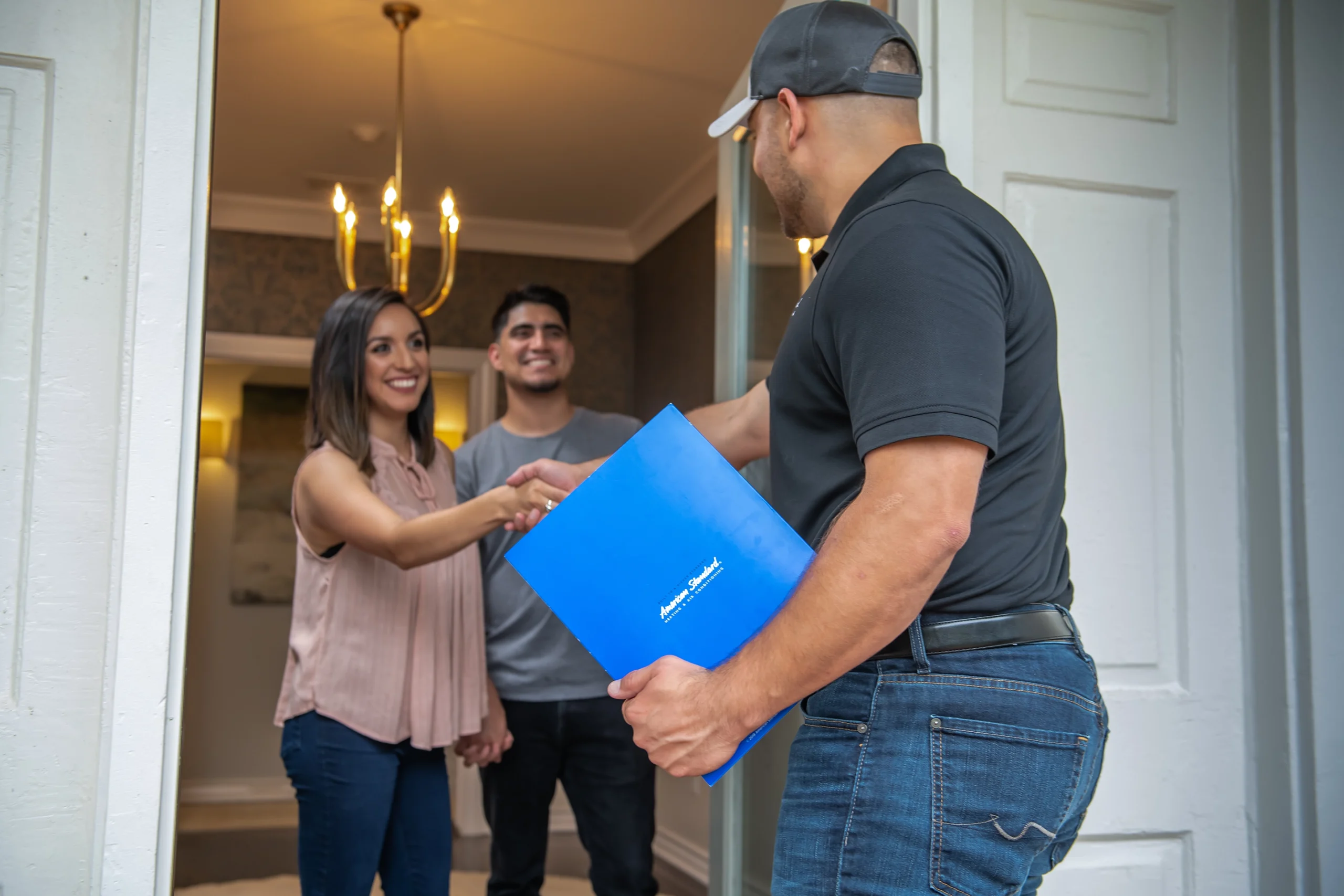an HVAC tech greets a couple at their front door. He's holding a folder.